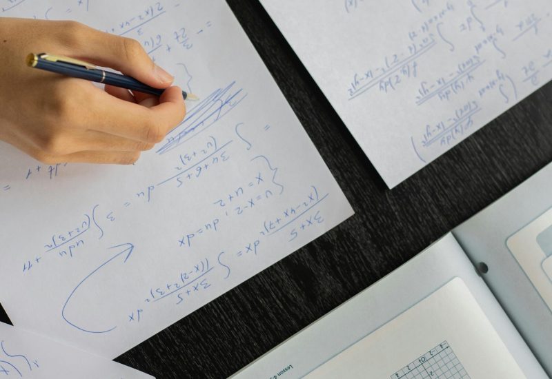 Close-up of a student solving mathematical equations with papers and textbook on a table.